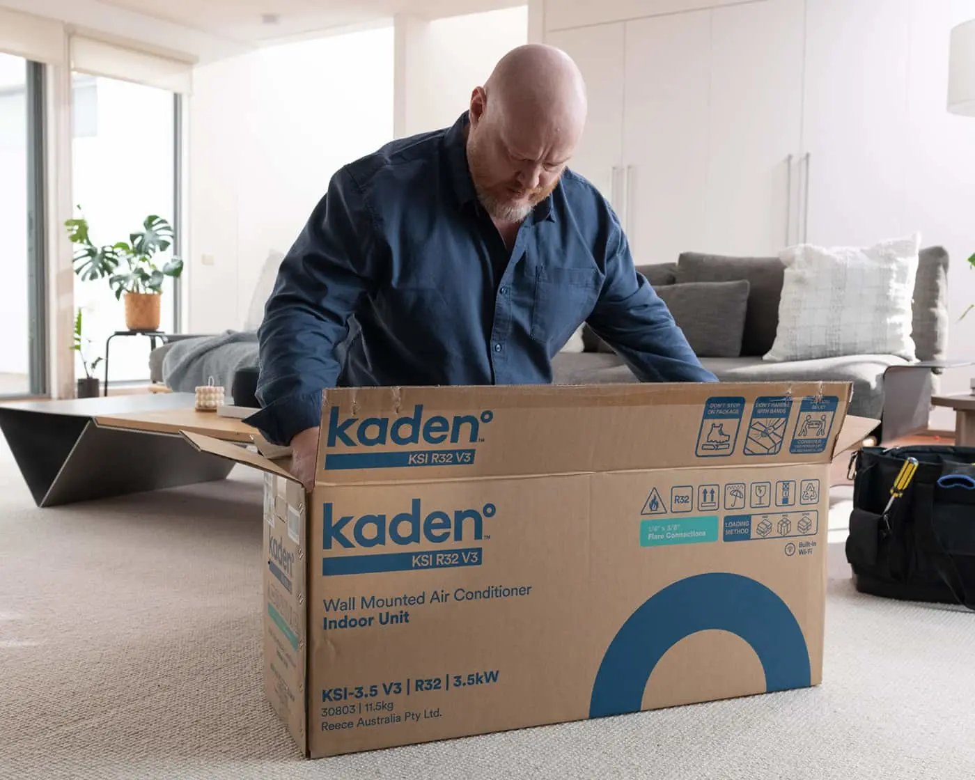 Man in blue shirt opening a Kaden air conditioner box in a bright living room with gray couch and plants.