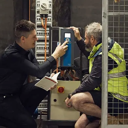 Two men inspecting an electrical control panel in an industrial setting, one holding a tablet and notebook.
