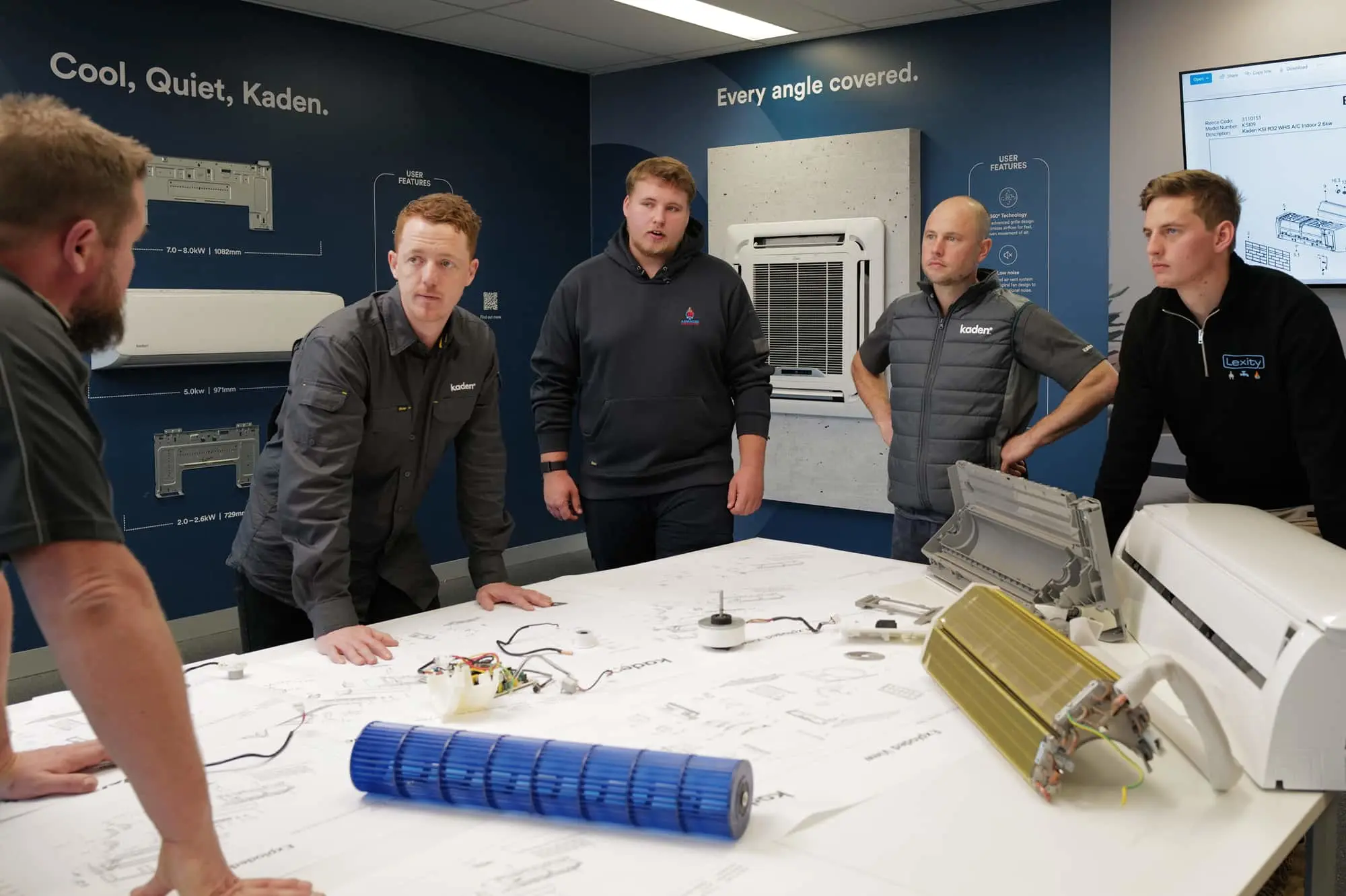 Five men in a meeting room examining air conditioning unit components on a table with a wall-mounted unit behind them.
