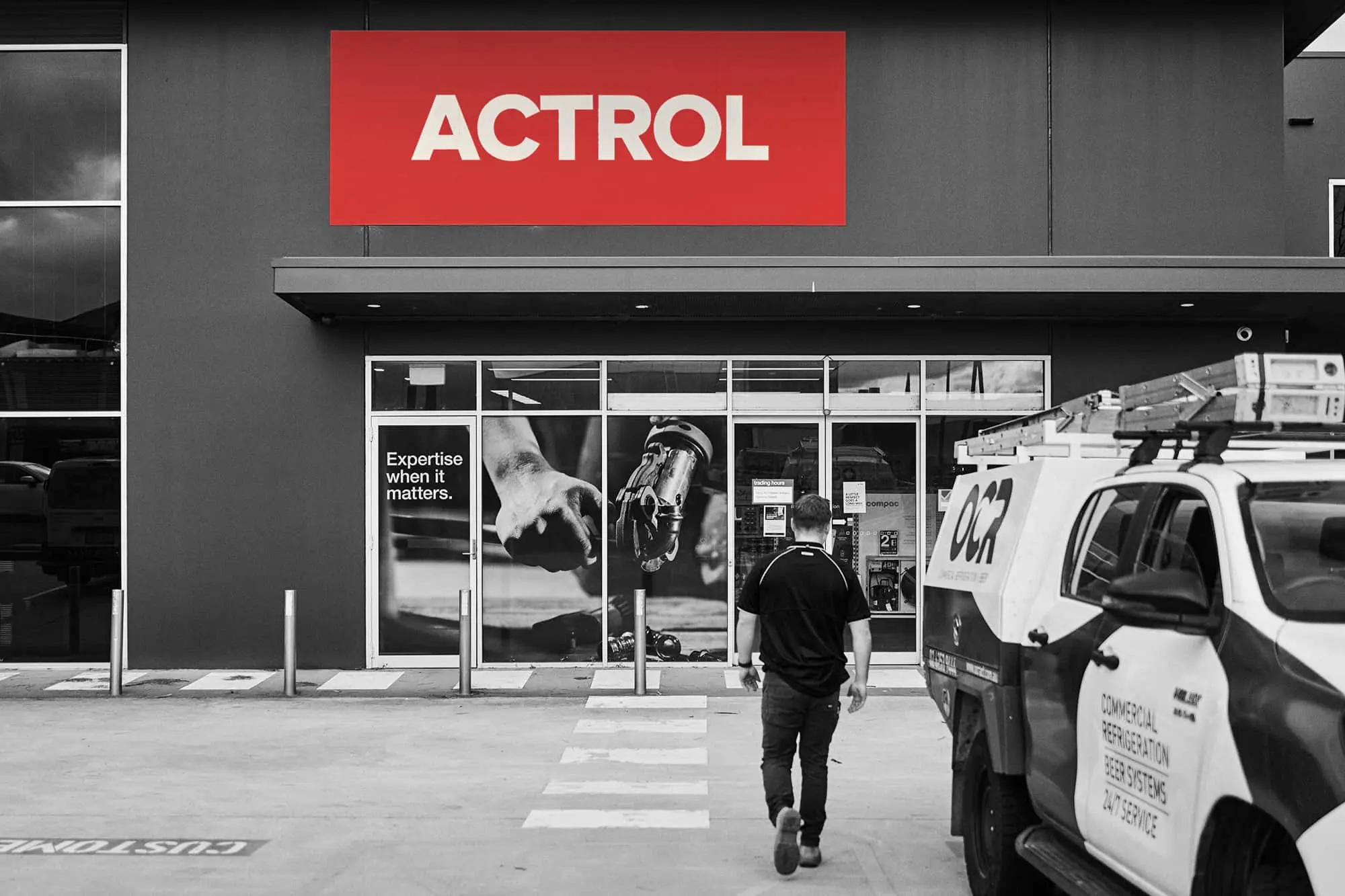 Person walking toward ACTROL storefront with red sign and glass doors, white van parked on right side.