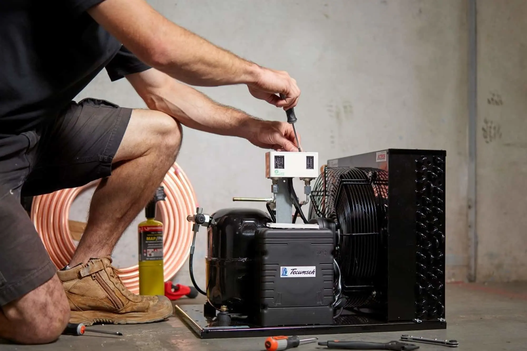 Person in black shirt and work boots using a screwdriver to work on a black refrigeration compressor unit.