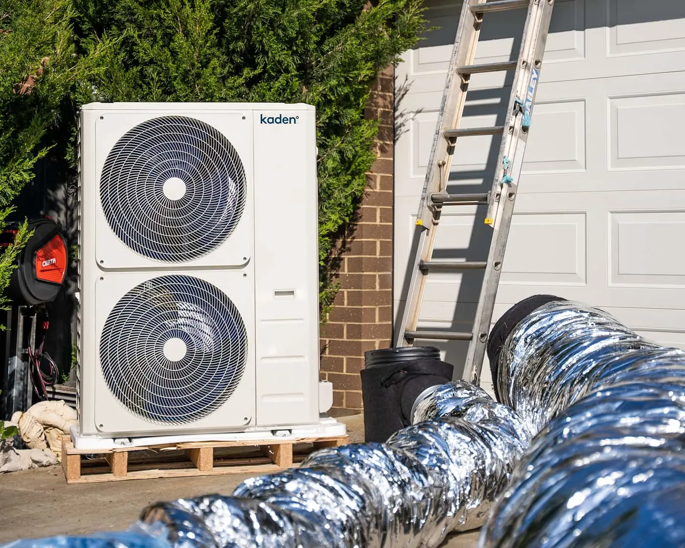 White HVAC unit with dual fans on wooden pallet beside brick wall, with ladder against garage door and silver ductwork.