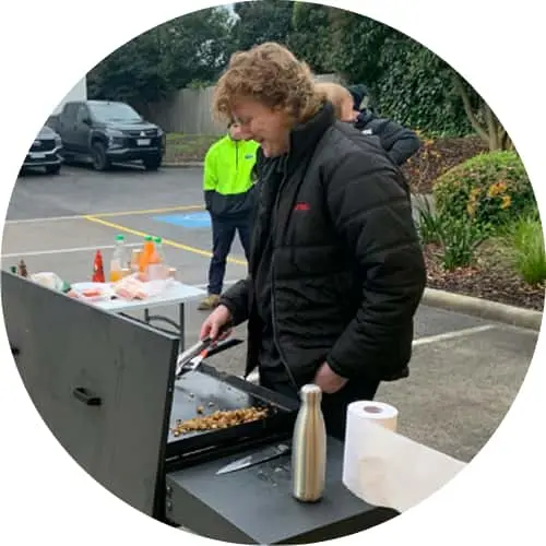 Person in black jacket cooking food on outdoor grill with another person in safety vest visible in background