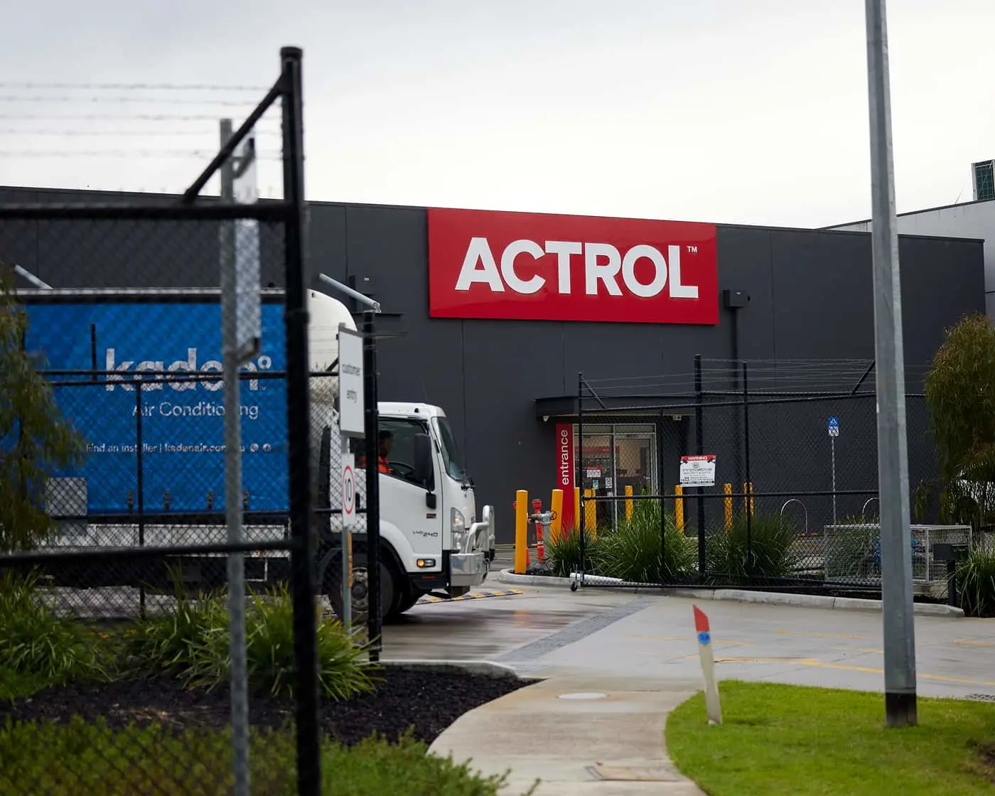 Dark gray industrial building with large red ACTROL sign, white truck at entrance, chain-link fence in foreground.