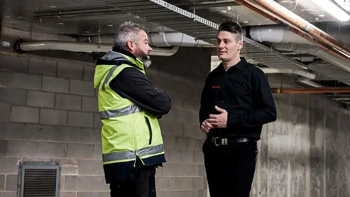 Two men in conversation in an unfinished building with exposed ceiling pipes and concrete block walls.