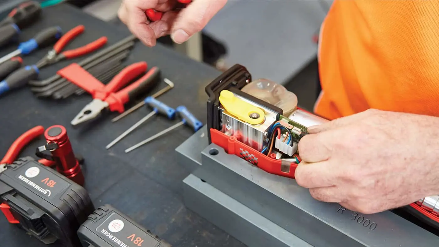 Person in orange shirt repairing an electronic device with tools and battery packs visible on workbench.
