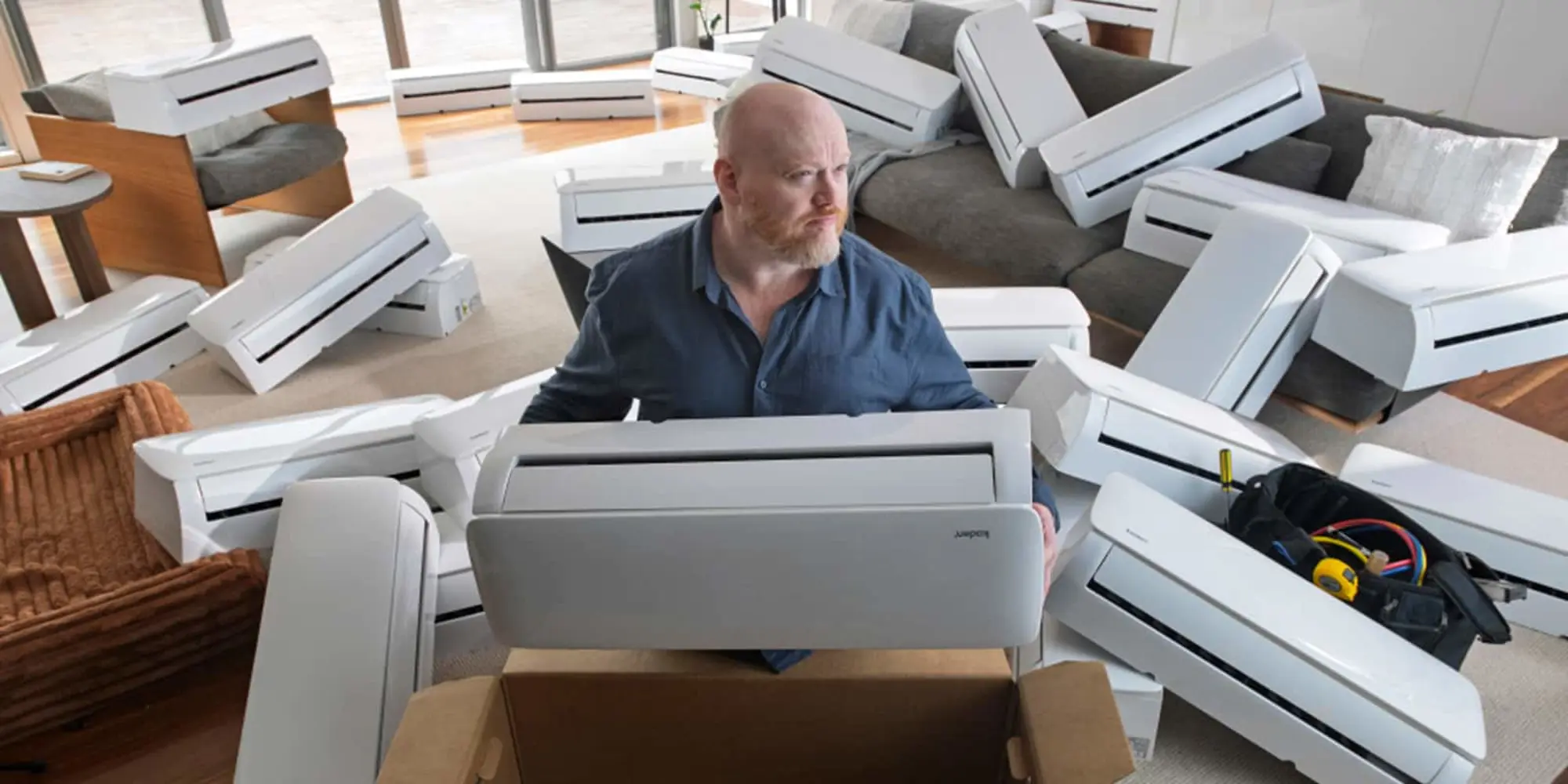 Man in blue shirt surrounded by multiple white air conditioning units in a living room with gray furniture.