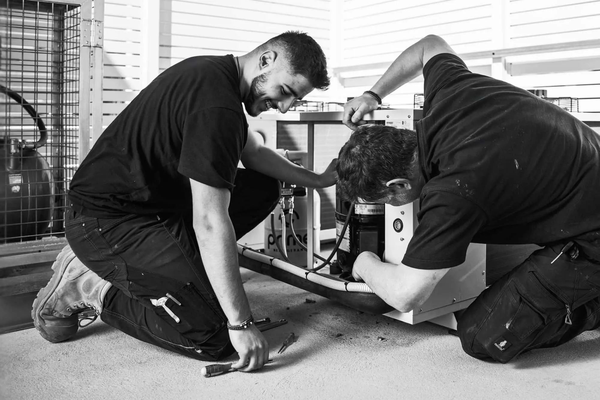 Two technicians in black shirts kneeling on floor repairing HVAC equipment in black and white photo