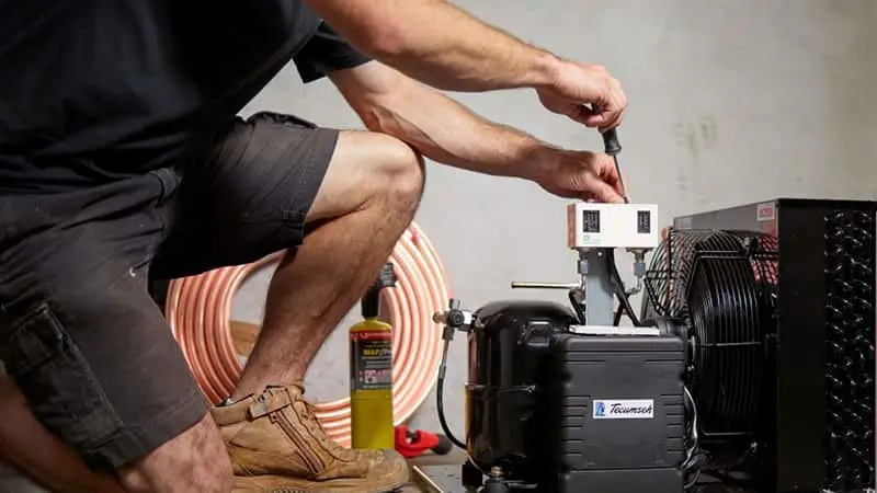 A technician kneeling beside an air conditioning unit, working on the compressor with copper coils visible nearby.