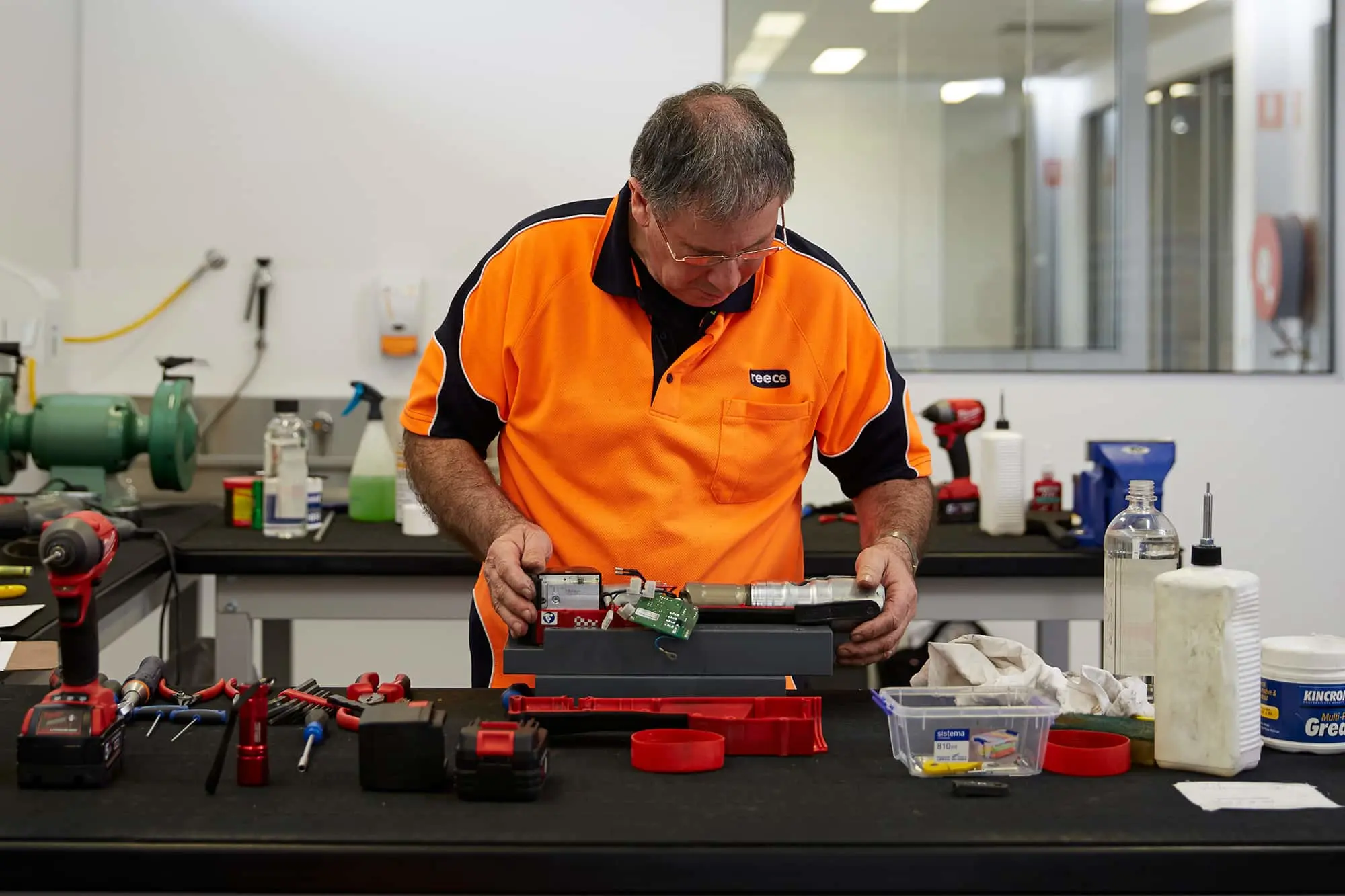Technician in orange and black uniform working on electronic equipment at a workbench with tools and supplies.