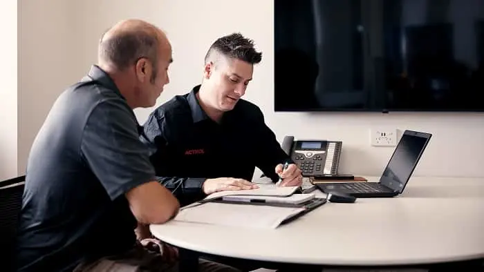 Two men in dark uniforms sit at a table reviewing documents, with a laptop, phone, and wall-mounted TV visible.