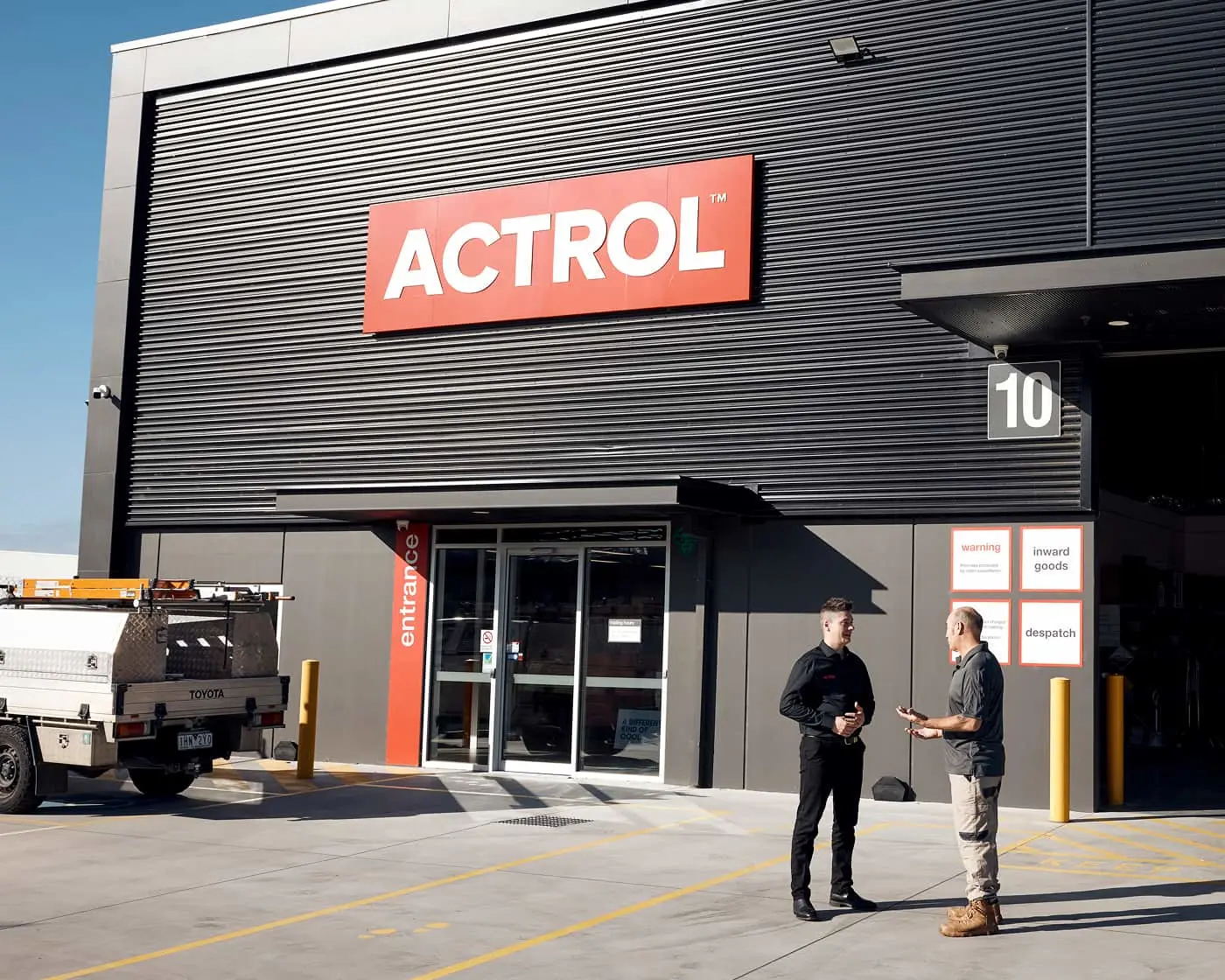 Two people talking outside an ACTROL warehouse building with a white truck parked nearby and number 10 on the wall.