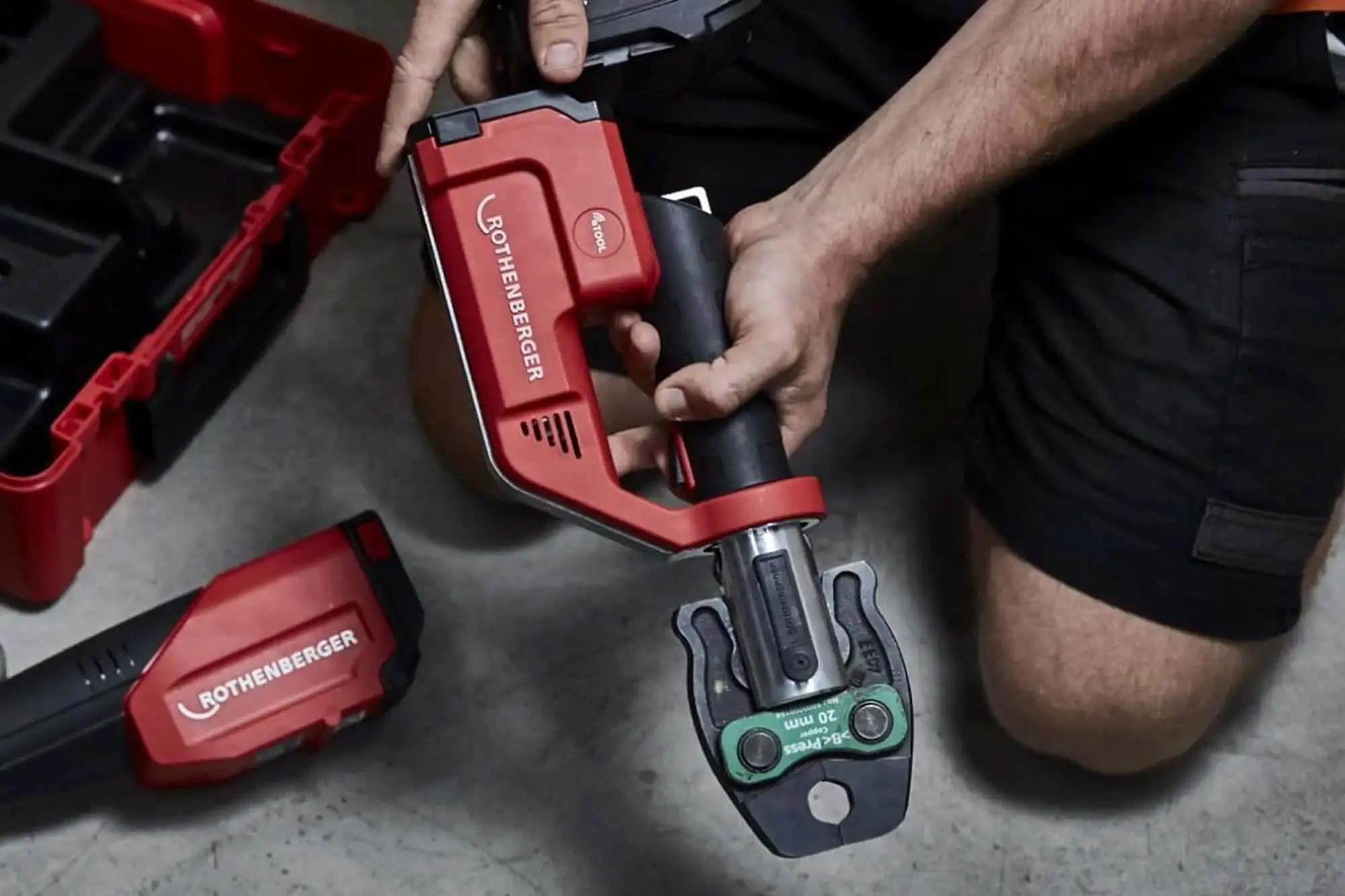 Hands holding a red Rothenberger cordless pressing tool with battery pack and red toolbox nearby on floor.