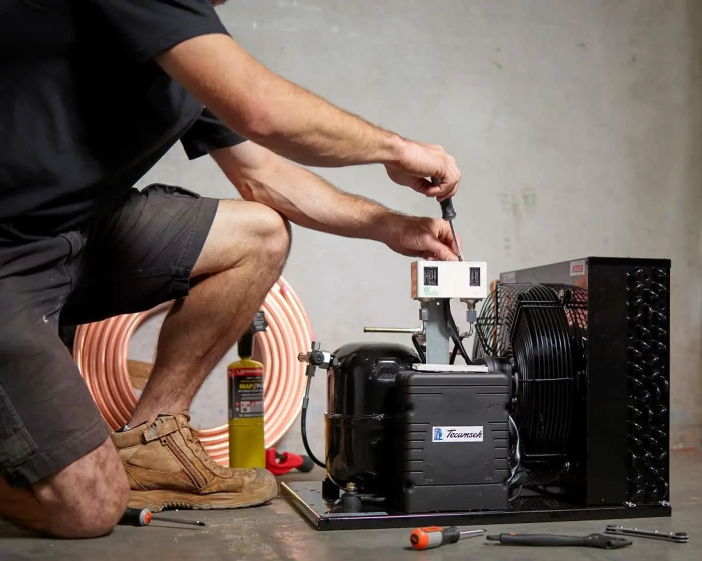 Person crouching while working on a refrigeration unit with copper tubing and tools on the floor.