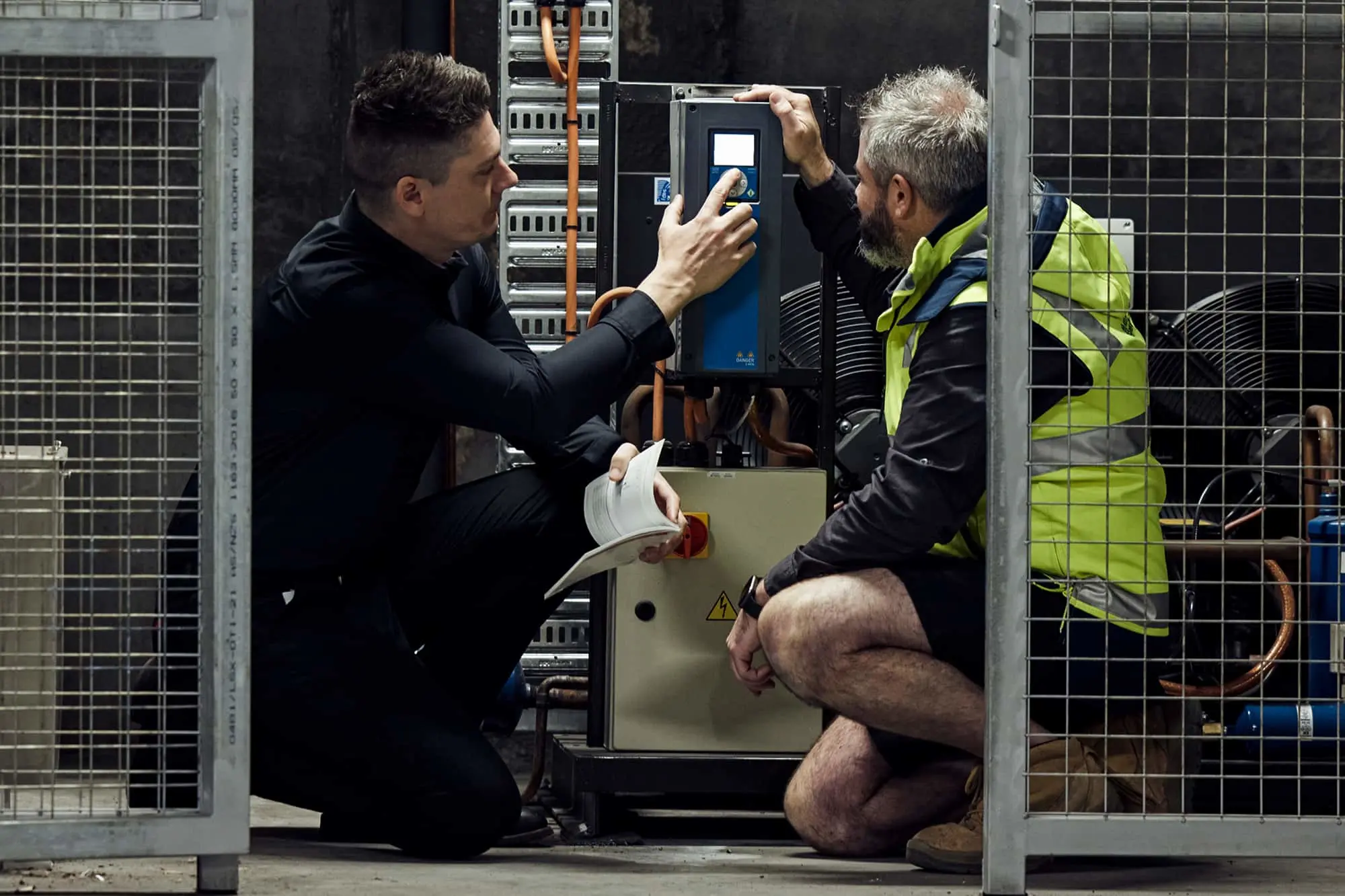 Two technicians working on industrial equipment with blue and black control panel in a facility with metal cages.