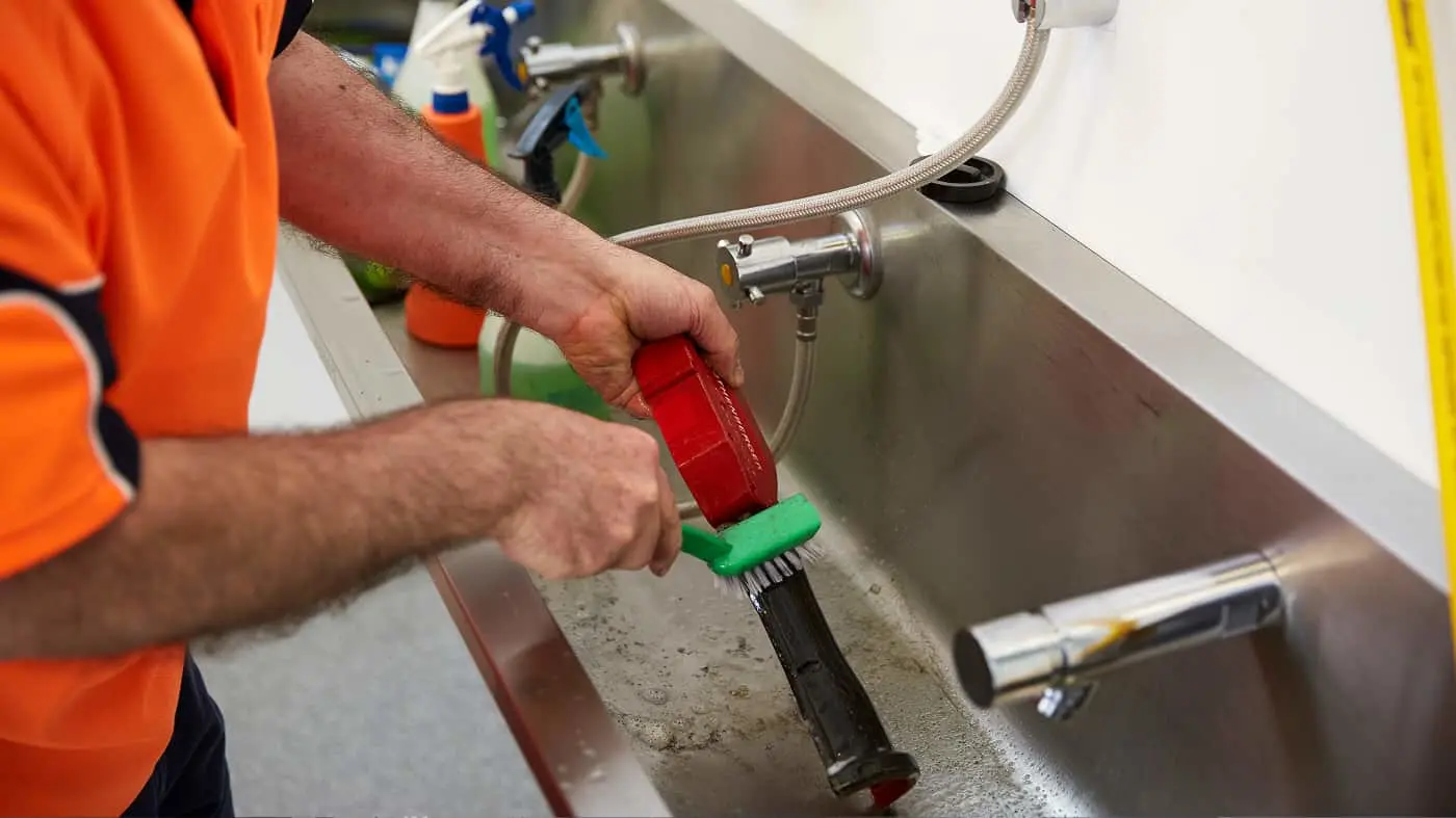 Plumber in orange shirt using a red and green brush tool to clean a kitchen sink drain, with cleaning supplies nearby.