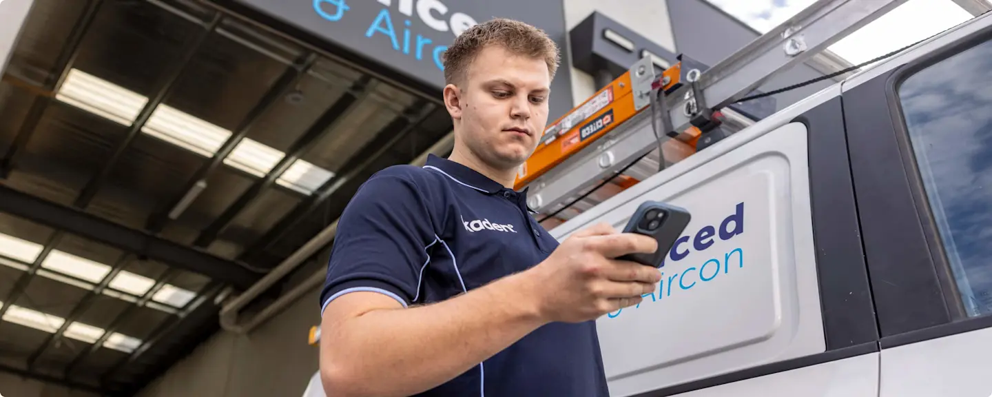 Man in navy uniform holding smartphone standing next to white service van with roof rack in warehouse setting.