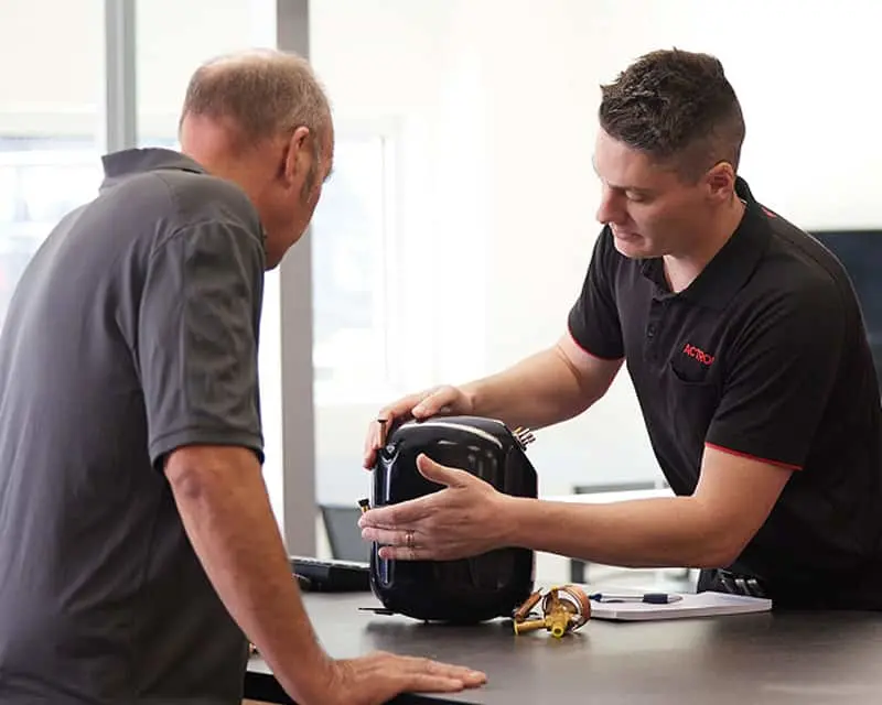 Two men examining a condensor on a counter, one in a gray shirt and one in a black polo shirt.