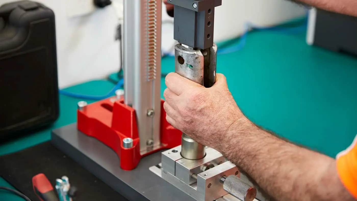 Hands operating a material testing machine with red and silver components on a turquoise surface in a laboratory setting.