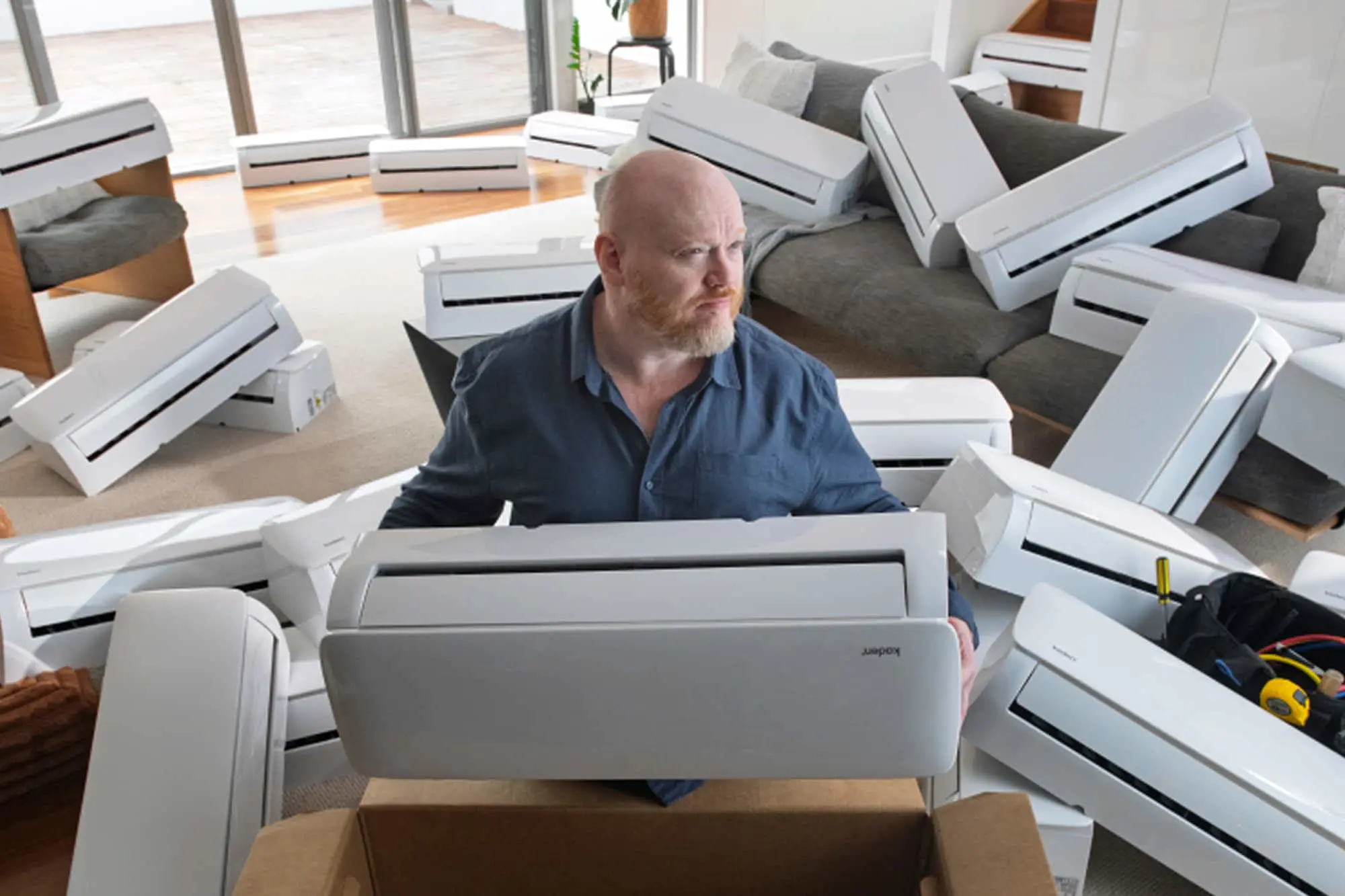Man in blue shirt surrounded by multiple white split system air conditioning units in a living room with gray couch and large windows.