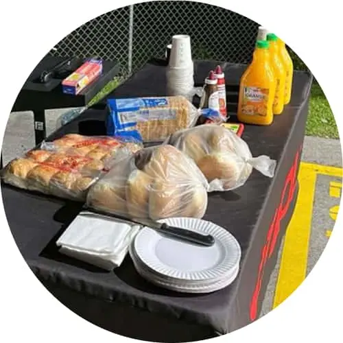 Picnic supplies and food items arranged on a bbq, including bagged bread, plates, cups, and condiments.