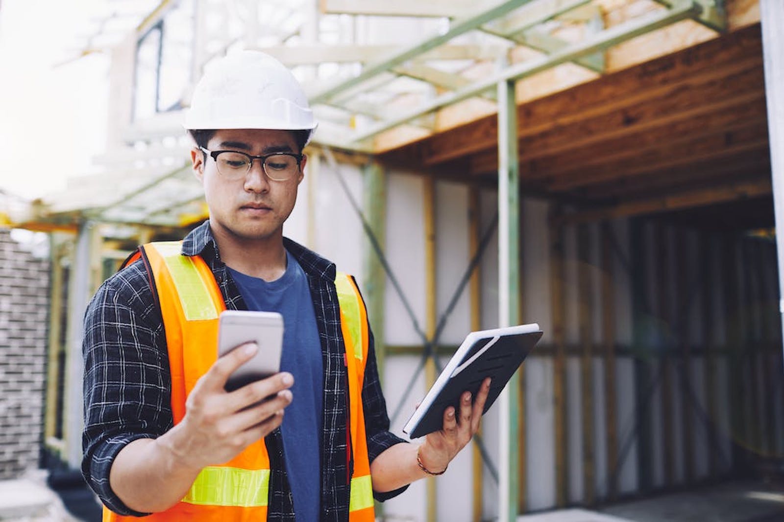 A worker in a safety vest and white hard hat holds a phone in one hand, and a notebook in another