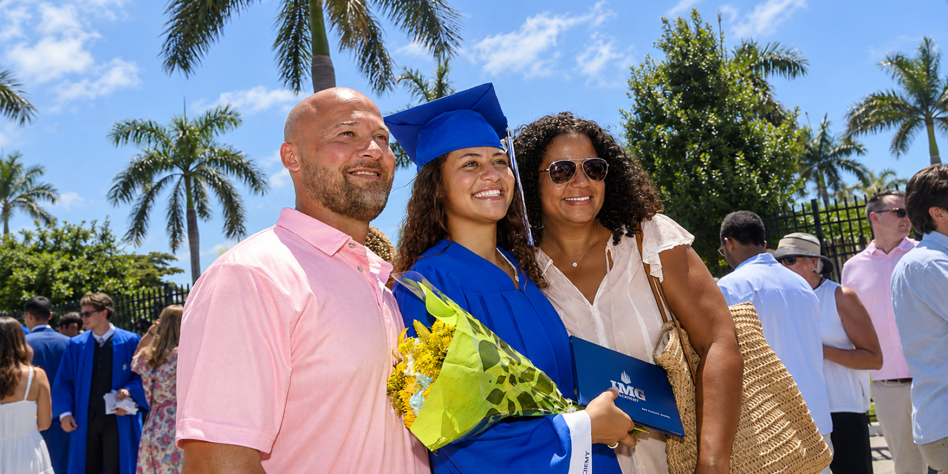 A graduate in a blue cap and gown poses with her family while holding flowers and a diploma folder.