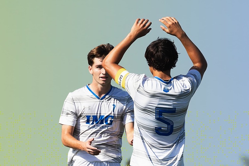 Two IMG Academy soccer players in white uniforms celebrate together on the field, raising their arms after a successful play under a clear sky.