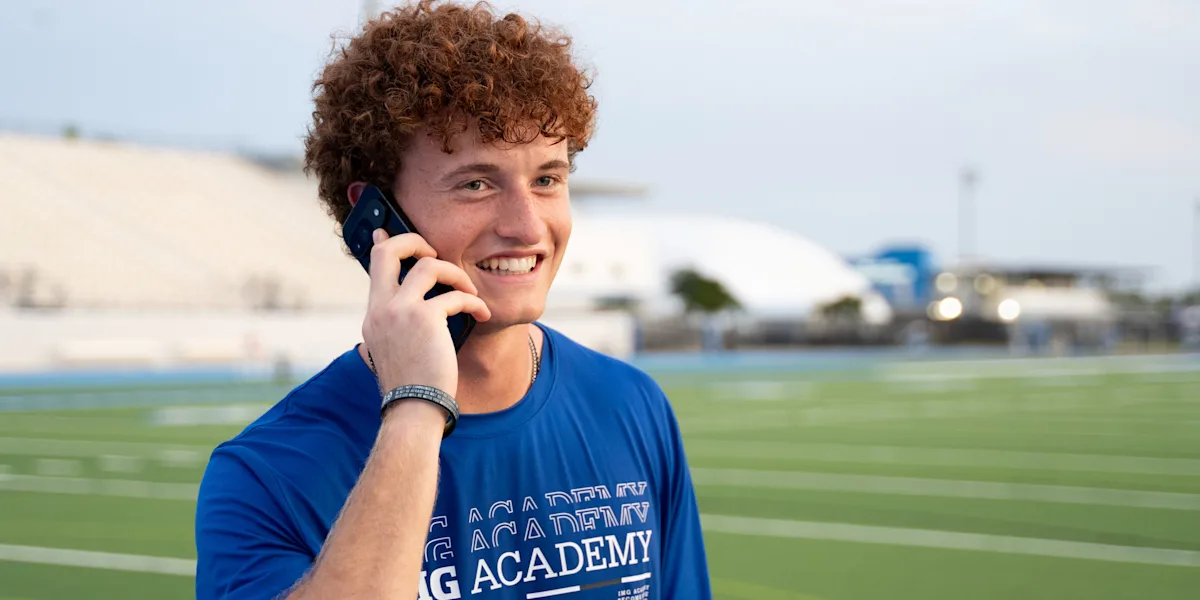 A smiling student-athlete talks on a phone beside a field.