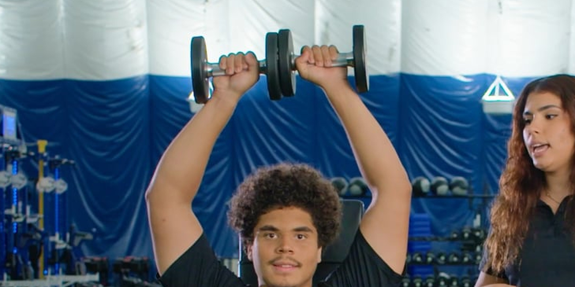 An athlete holds dumbbells overhead while a trainer observes.