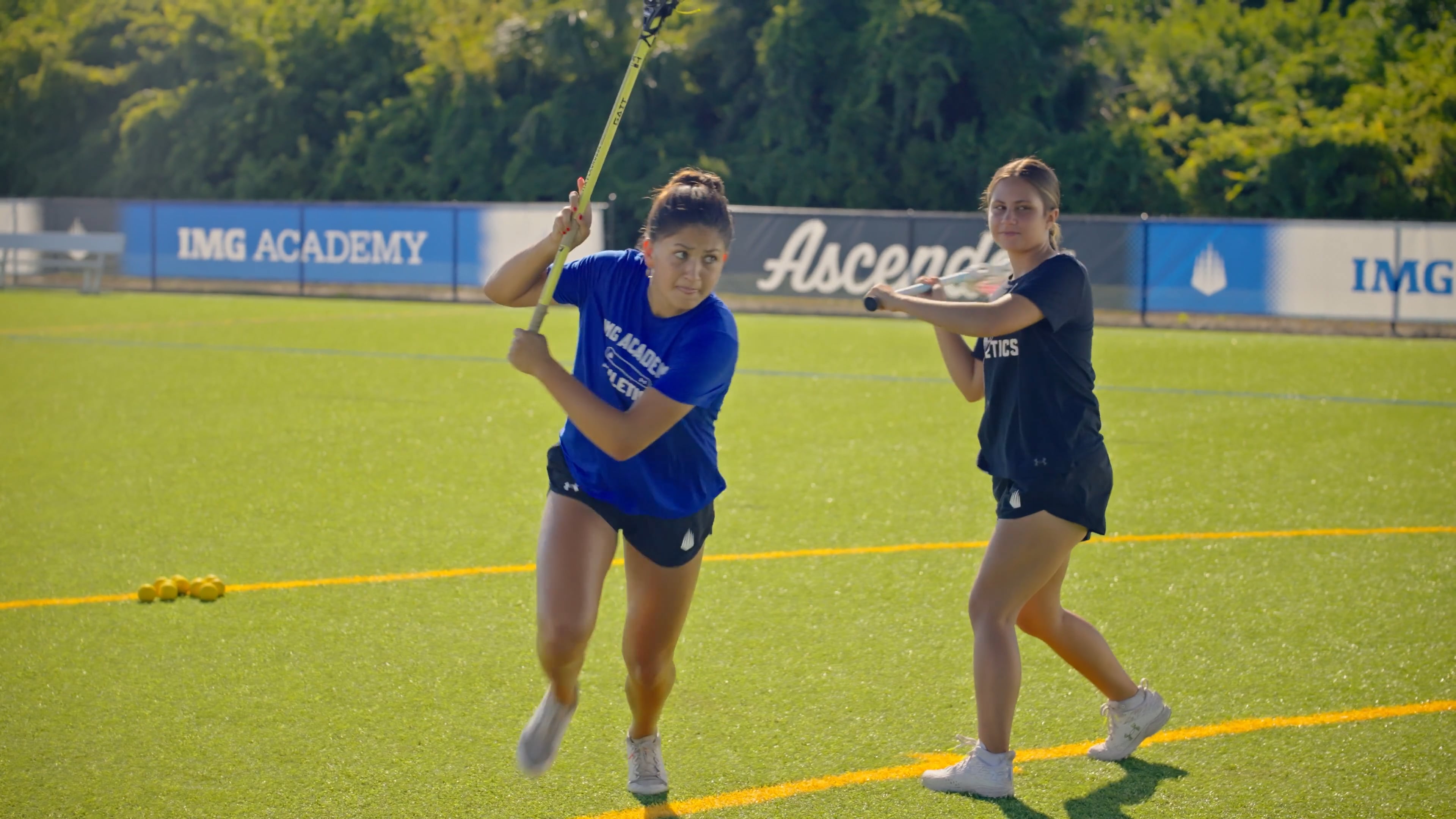 Two female lacrosse players practice on a green turf field under bright sunlight. The player in the foreground wears a blue shirt and black shorts, running forward with her lacrosse stick raised.