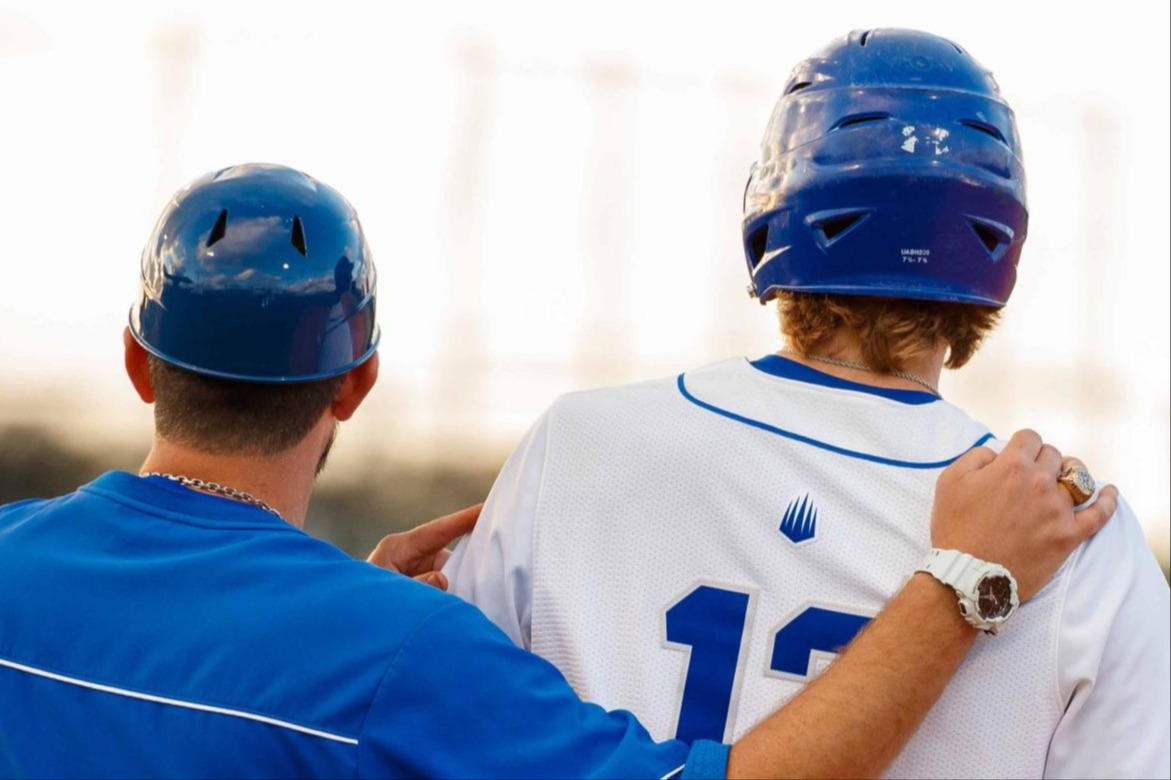 A baseball coach with his arm around a player's shoulder, viewed from behind, during a game at sunset.