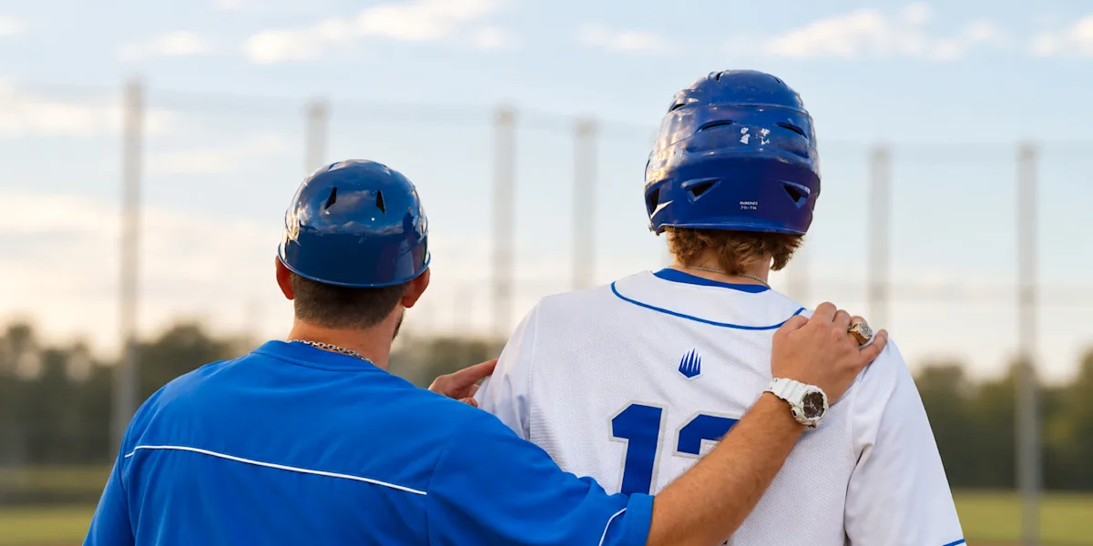A baseball coach rests a hand on a helmeted player’s shoulder as they look across the field.
