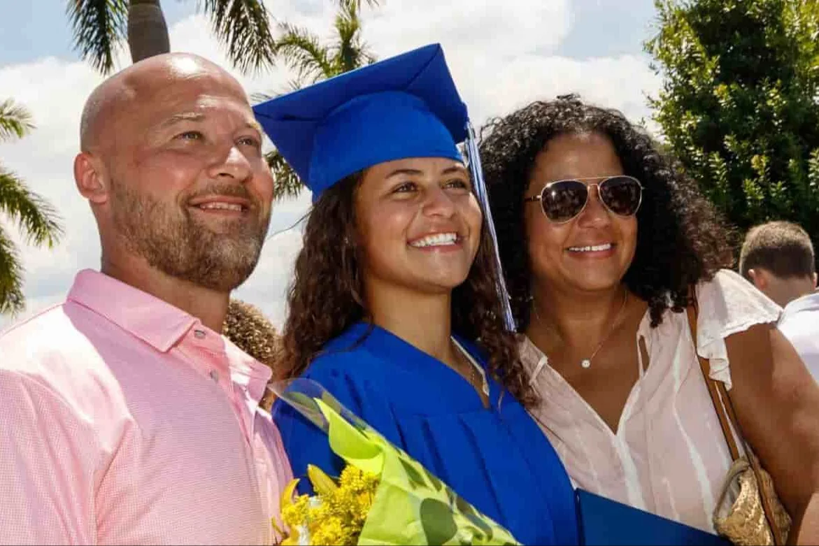 A young woman in a bright blue cap and gown poses with her family at an outdoor graduation ceremony, holding a bouquet of yellow flowers. Palm trees and other smiling attendees are visible in the sunny background.