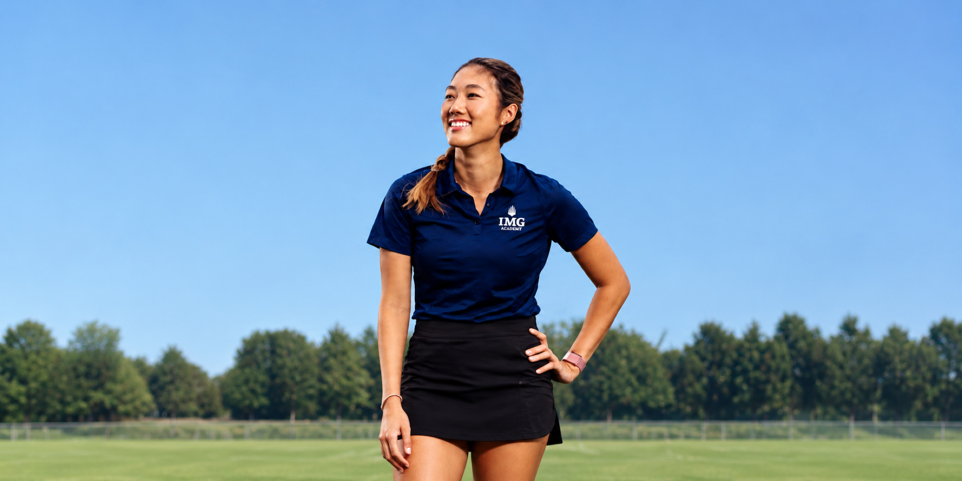 A smiling coach stands with one hand on her hip on a grass field.