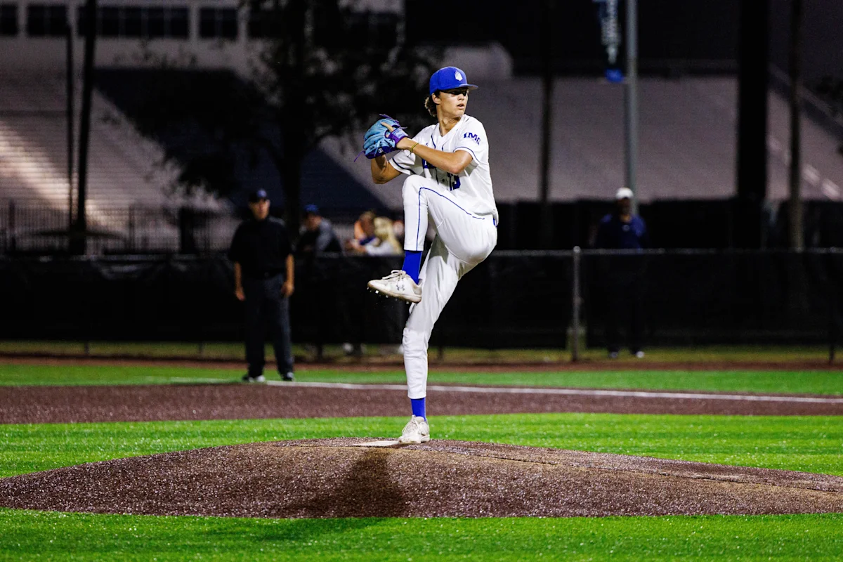 High school baseball player in full white uniform winds up to pitch on a lit field at night. He stands tall on the mound with focused posture, ready to deliver the ball.