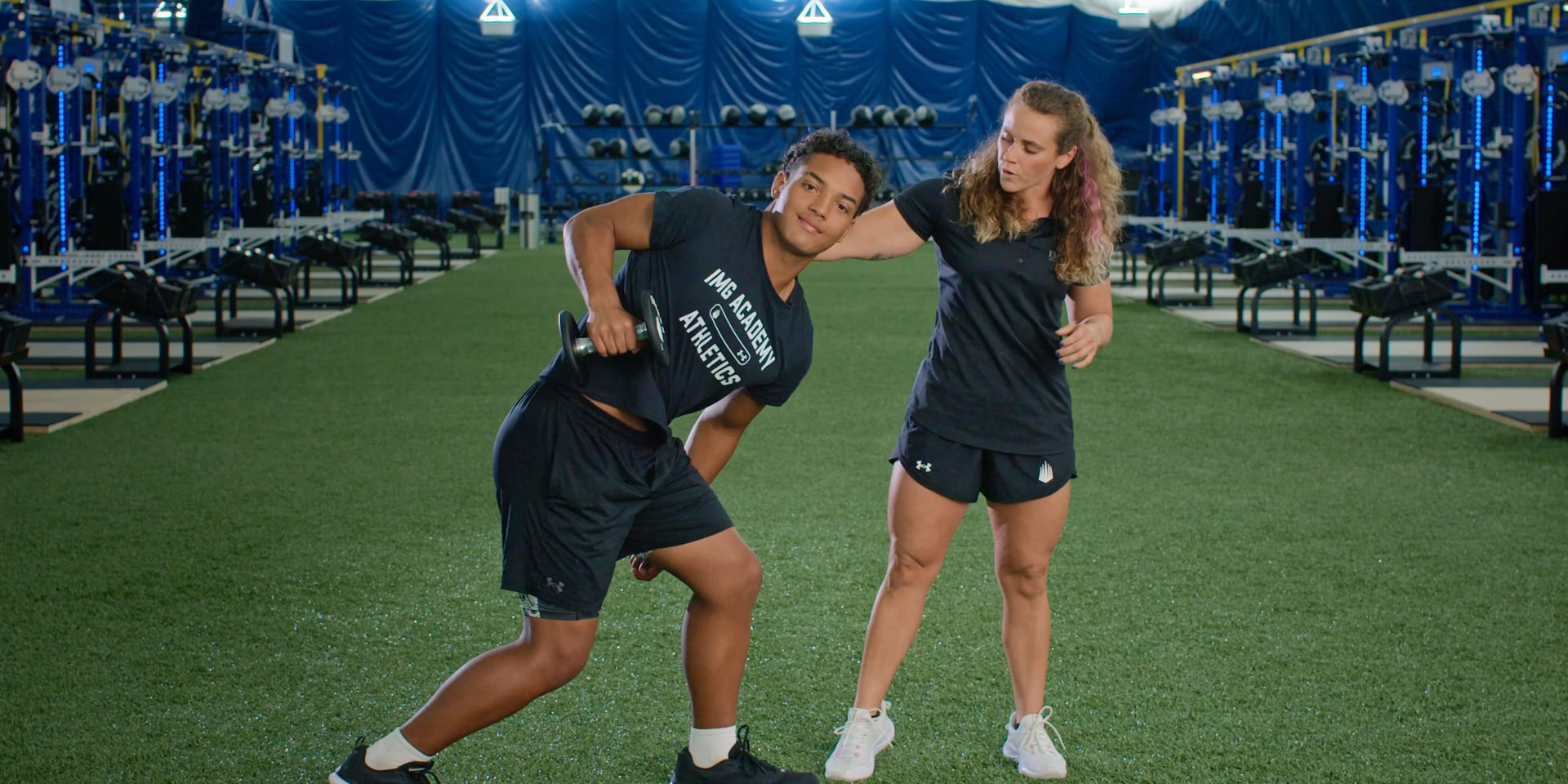 A trainer guides an athlete through a standing exercise on indoor turf.