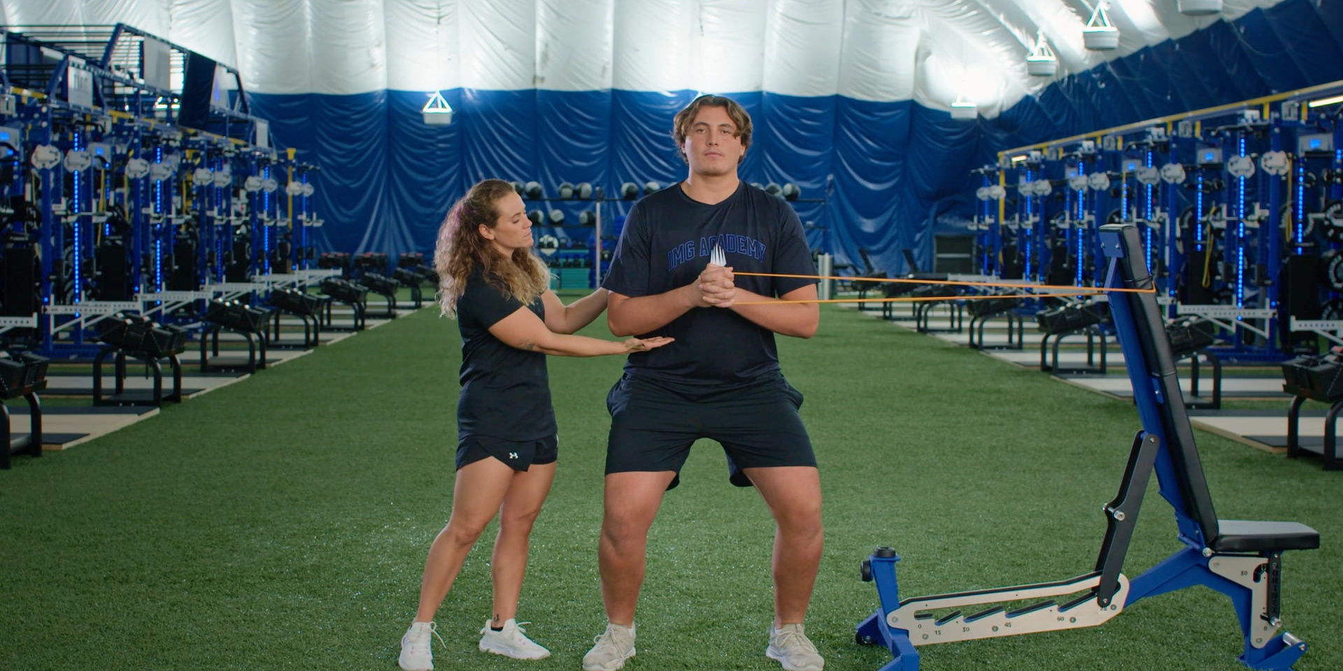 A trainer guides an athlete through a standing resistance-band exercise.
