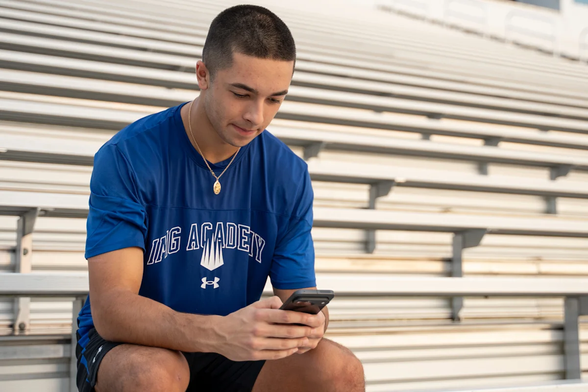Student-athlete sitting in bleachers and using a smartphone, representing how athletes can engage with college recruiters through social media.