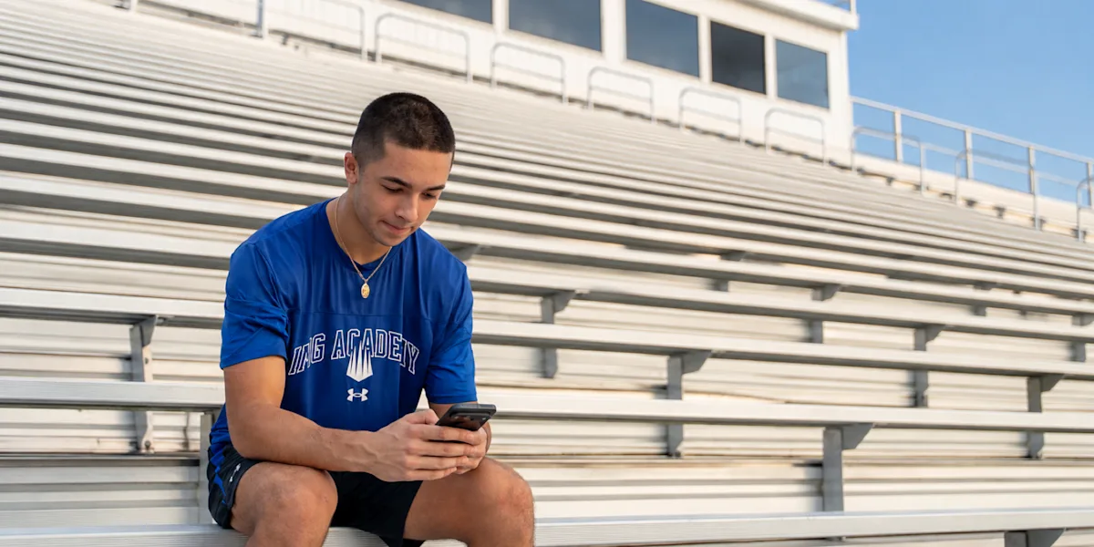 A student-athlete looks at his phone while seated on outdoor bleachers.