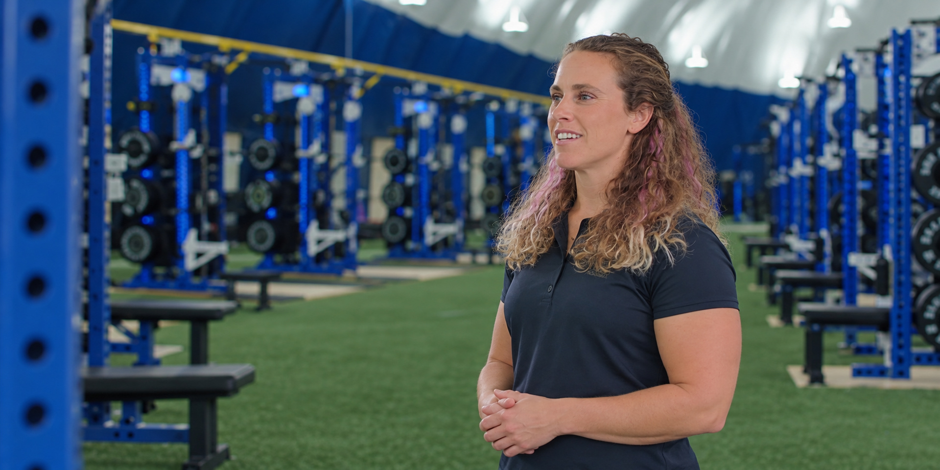 A trainer stands beside weight racks with her hands clasped.