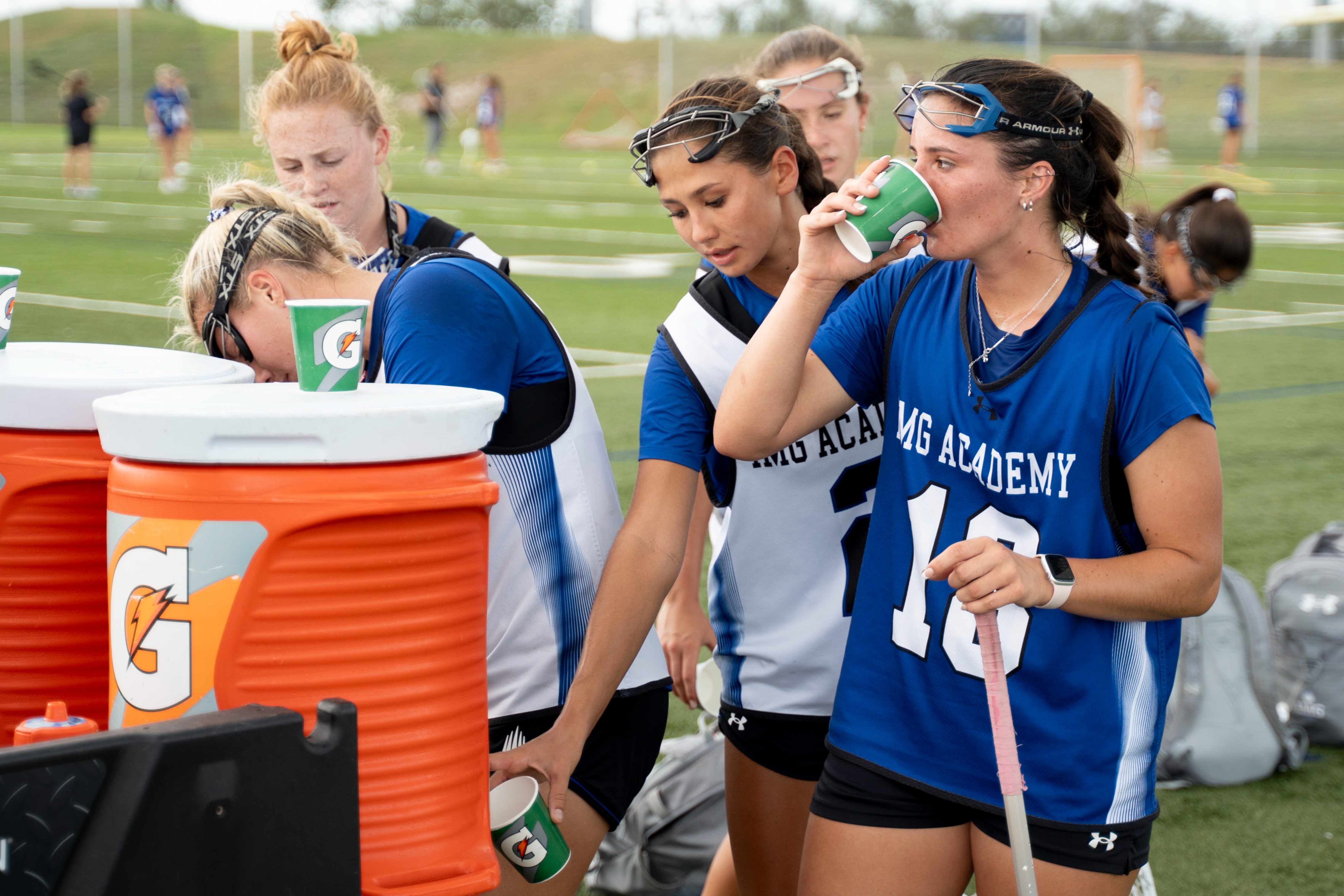 Female lacrosse athletes taking a hydration break, drinking from cups near orange Gatorade coolers on the sideline of a field.