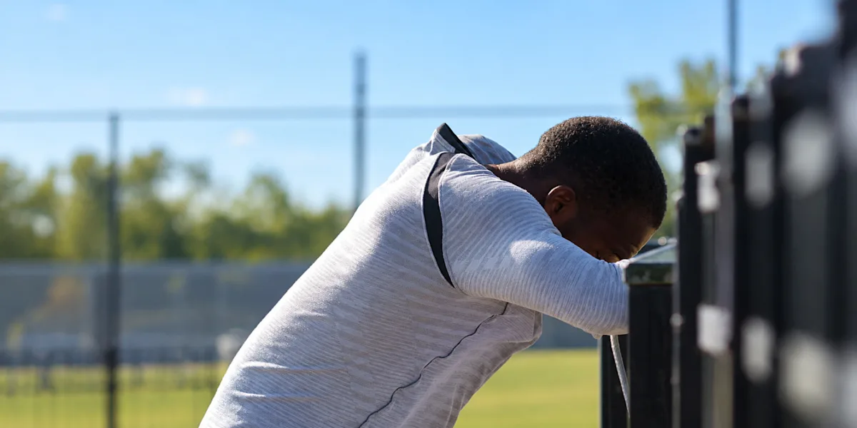 An athlete leans against a fence with his head resting on his arms.