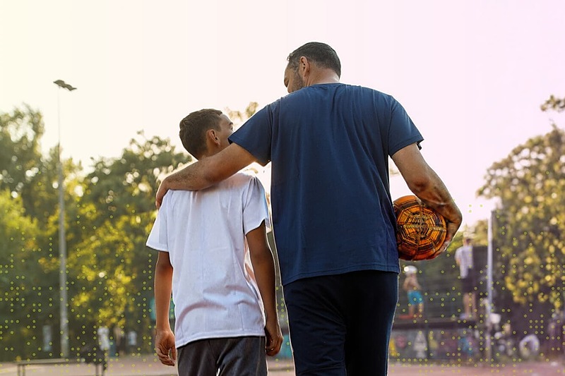 Parent walks with a young athlete on an outdoor sports field, arm around the athlete’s shoulders while holding a soccer ball, symbolizing mentorship, guidance, and support.