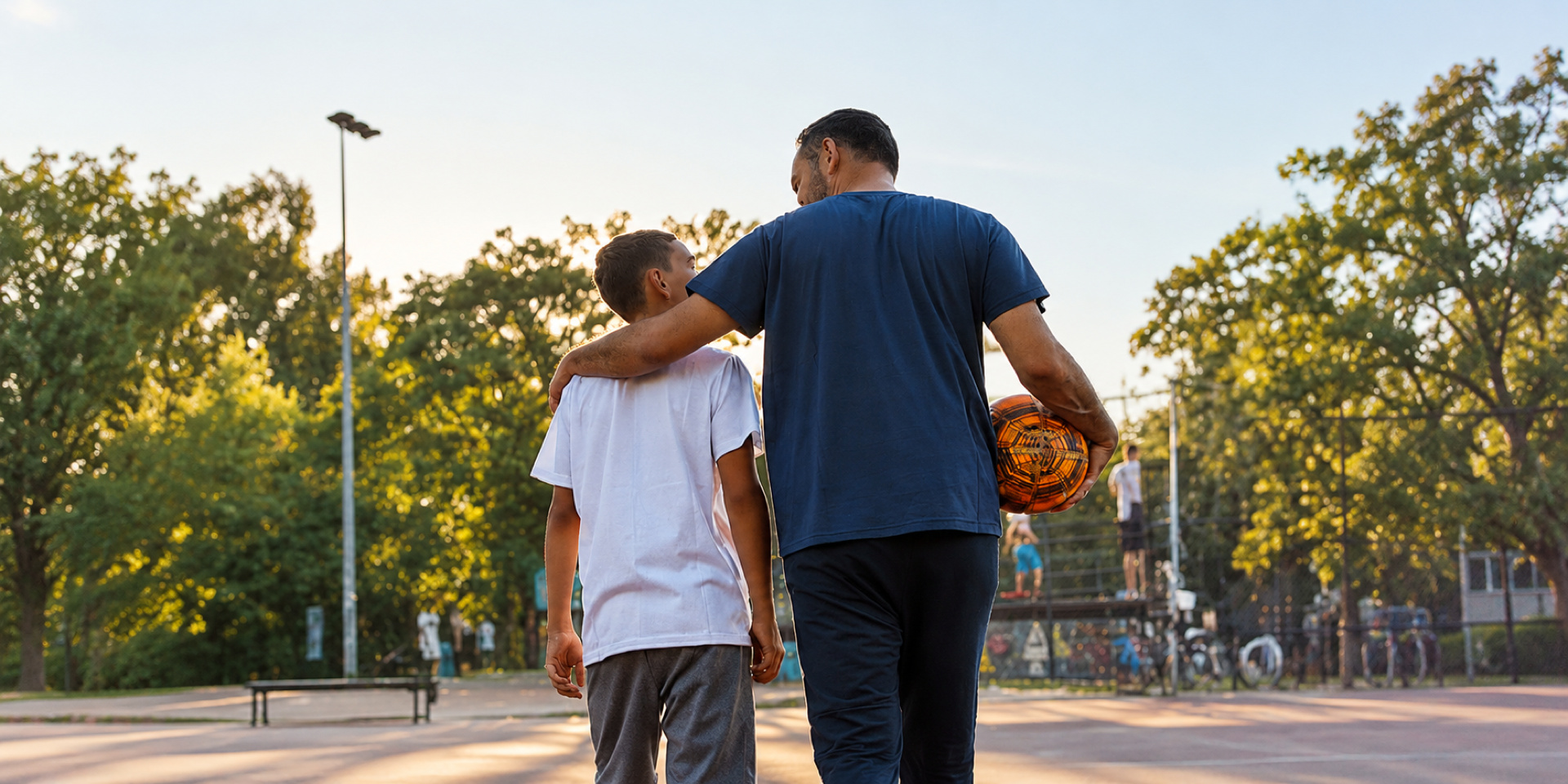An adult carrying a basketball walks with an arm around a young athlete on an outdoor court.