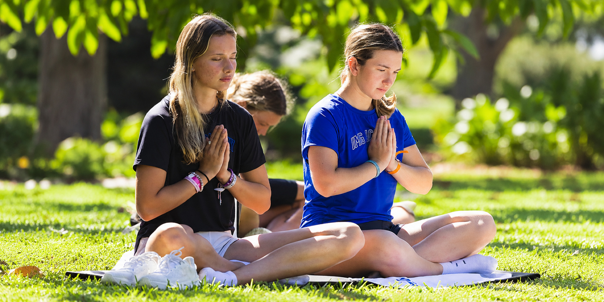 Students sit cross-legged on yoga mats outdoors with their hands together in front of their chests.