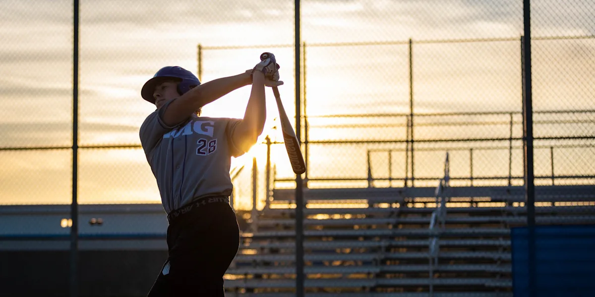 A baseball player follows through on a swing in evening sunlight.