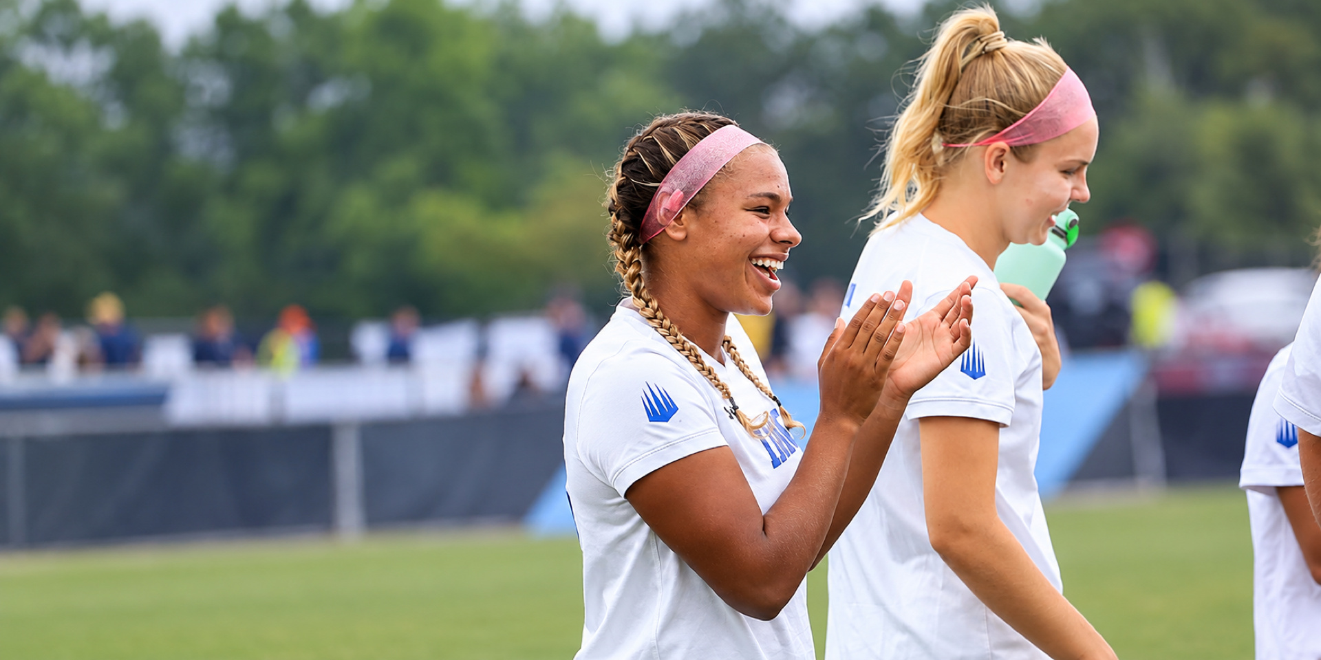 Two soccer players walk across a field, with one smiling and applauding.