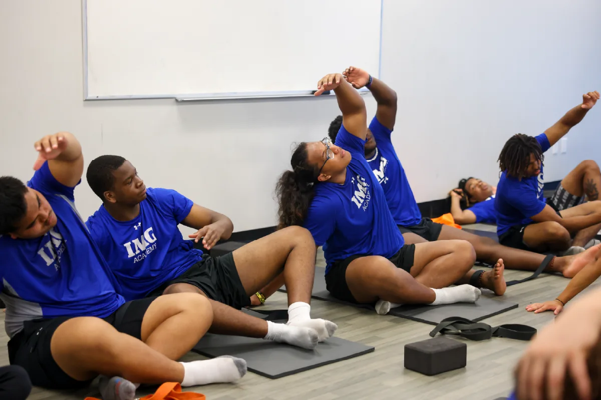 Athletes sitting on fitness mats inside a classroom performing a seated overhead side stretch during a mobility routine.