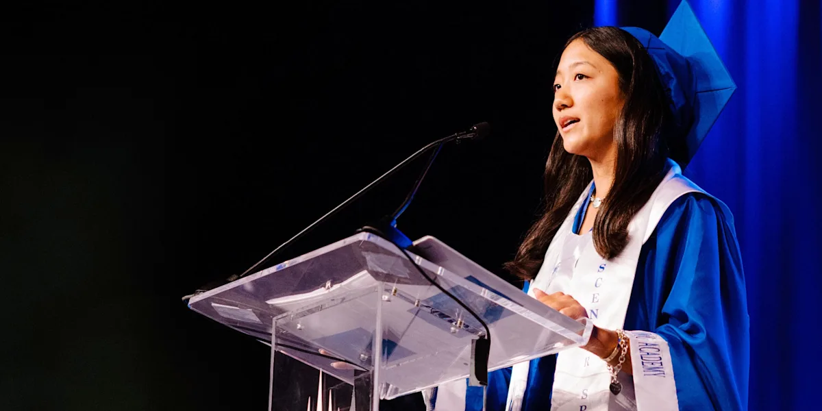 A graduate in a blue cap and gown speaks at a podium.
